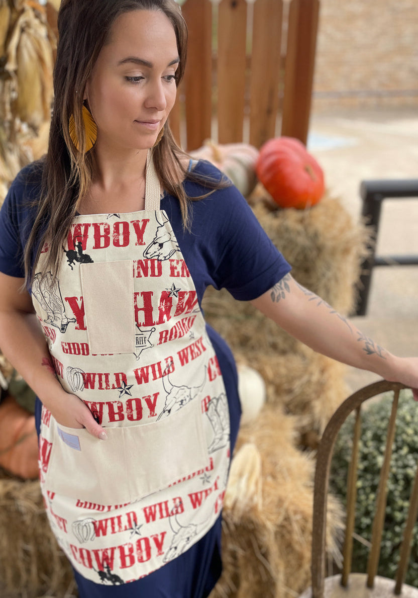 Woman wearing a cowboy-themed apron outdoors with hay bales and pumpkins in the background