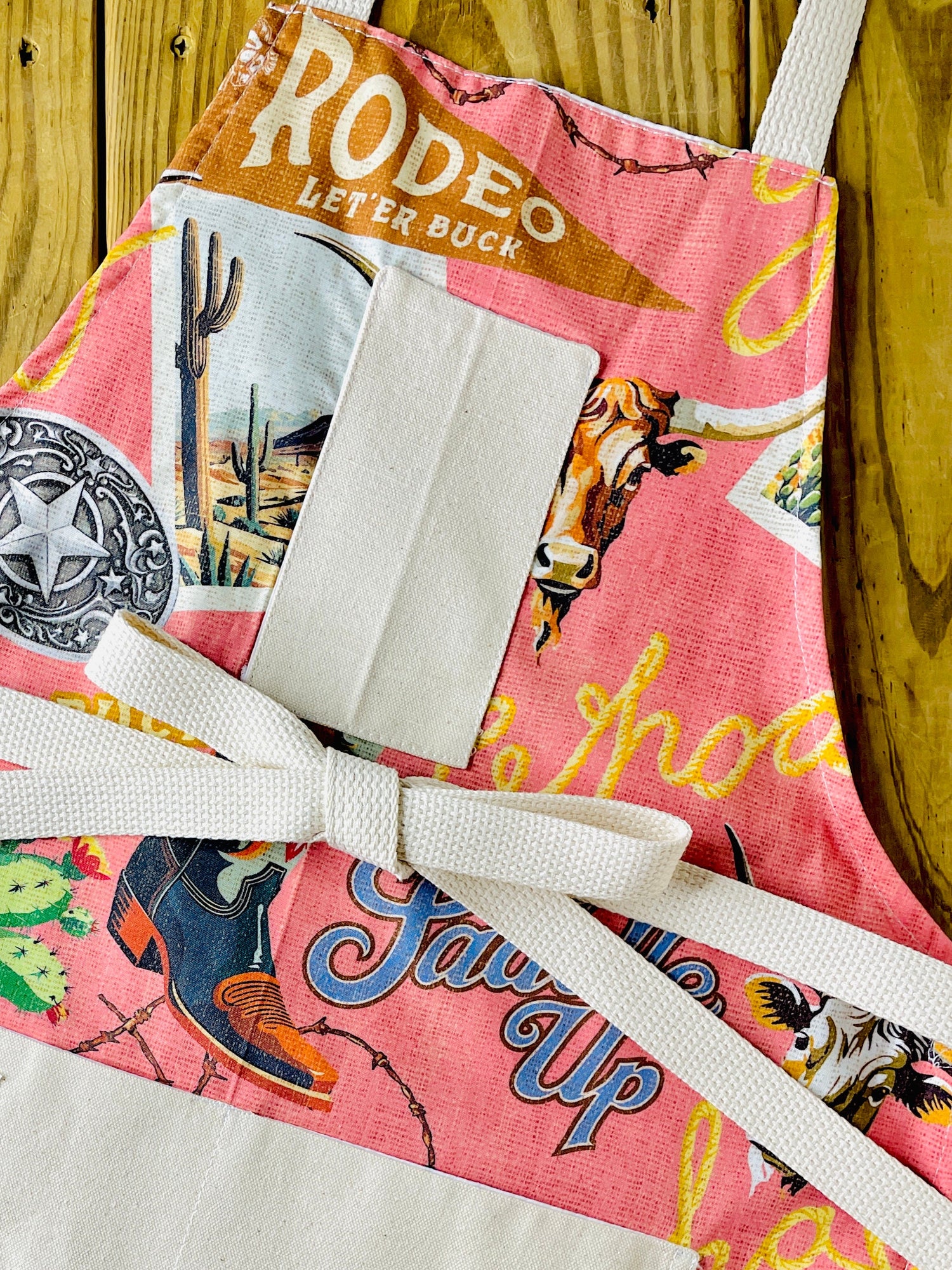 Amanda Bright Design colorful cotton apron with cowboy-themed design let ‘er buck saddle up buckle rodeo cowboy boot cactus postcard western