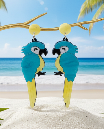 Amanda Bright Designs Blue and yellow parrot acrylic earrings on a branch with a beach background