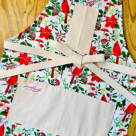 Amanda Bright Designs festive Christmas floral print apron featuring red cardinals, poinsettias, holly leaves, pinecones, and red mushrooms
