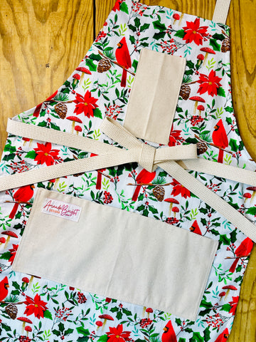 Amanda Bright Designs festive Christmas floral print apron featuring red cardinals, poinsettias, holly leaves, pinecones, and red mushrooms