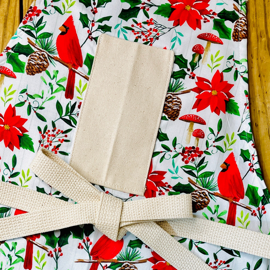 Amanda Bright Designs festive Christmas print apron featuring red cardinals, poinsettias, holly leaves, pinecones, and red mushrooms