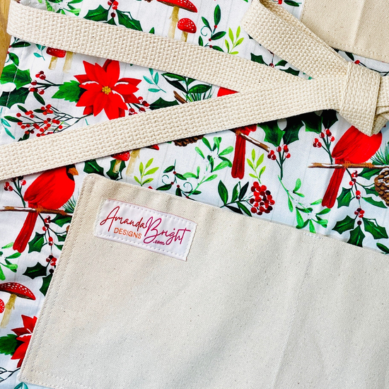 Amanda Bright Designs festive Christmas floral print apron featuring red cardinals, poinsettias, holly leaves, pinecones, and red mushrooms