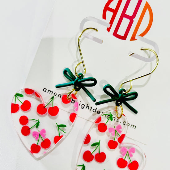 Amanda Bright Designs pair of heart-shaped acrylic dangle earrings with red cherries, green stems, and small pink bows, hanging from gold hooks with dark green ribbon bows. 