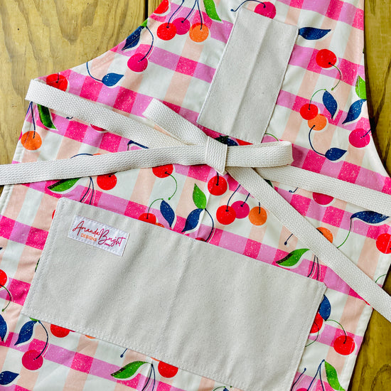 Amanda Bright Designs retro 1950s style cherries gingham plaid print apron in pink orange and cream with red cherries with modern vibe and navy and green leaves