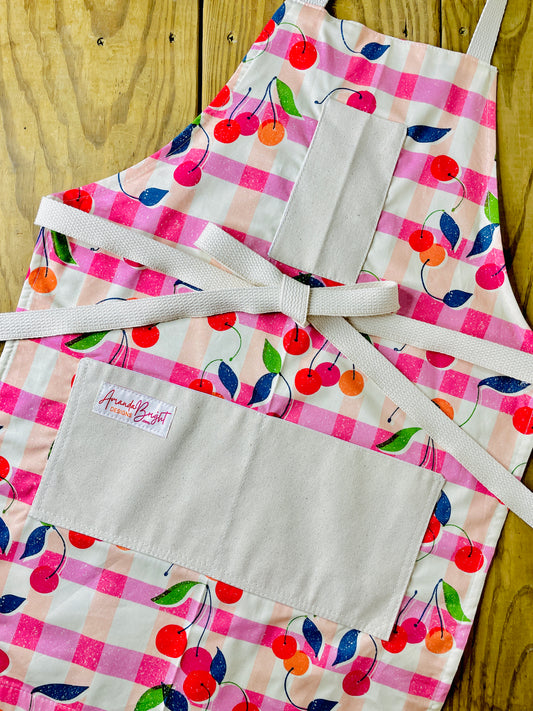 Amanda Bright Designs retro 1950s style cherries gingham plaid print apron in pink orange and cream with red cherries with modern vibe and navy and green leaves