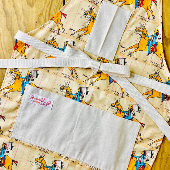 Amanda Bright Designs cheyenne warrior indian chief apron with cream background and a repeating print of indian on horseback wearing red and blue with his headdress on and figural marks on the pattern