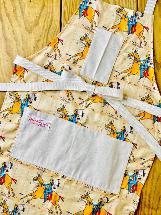 Amanda Bright Designs cheyenne warrior indian chief apron with cream background and a repeating print of indian on horseback wearing red and blue with his headdress on and figural marks on the pattern