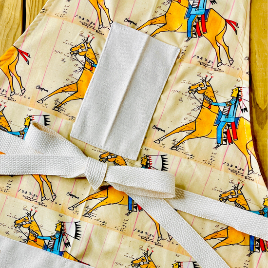 Amanda Bright Designs cheyenne warrior indian chief apron with cream background and a repeating print of indian on horseback wearing red and blue with his headdress on and figural marks on the pattern