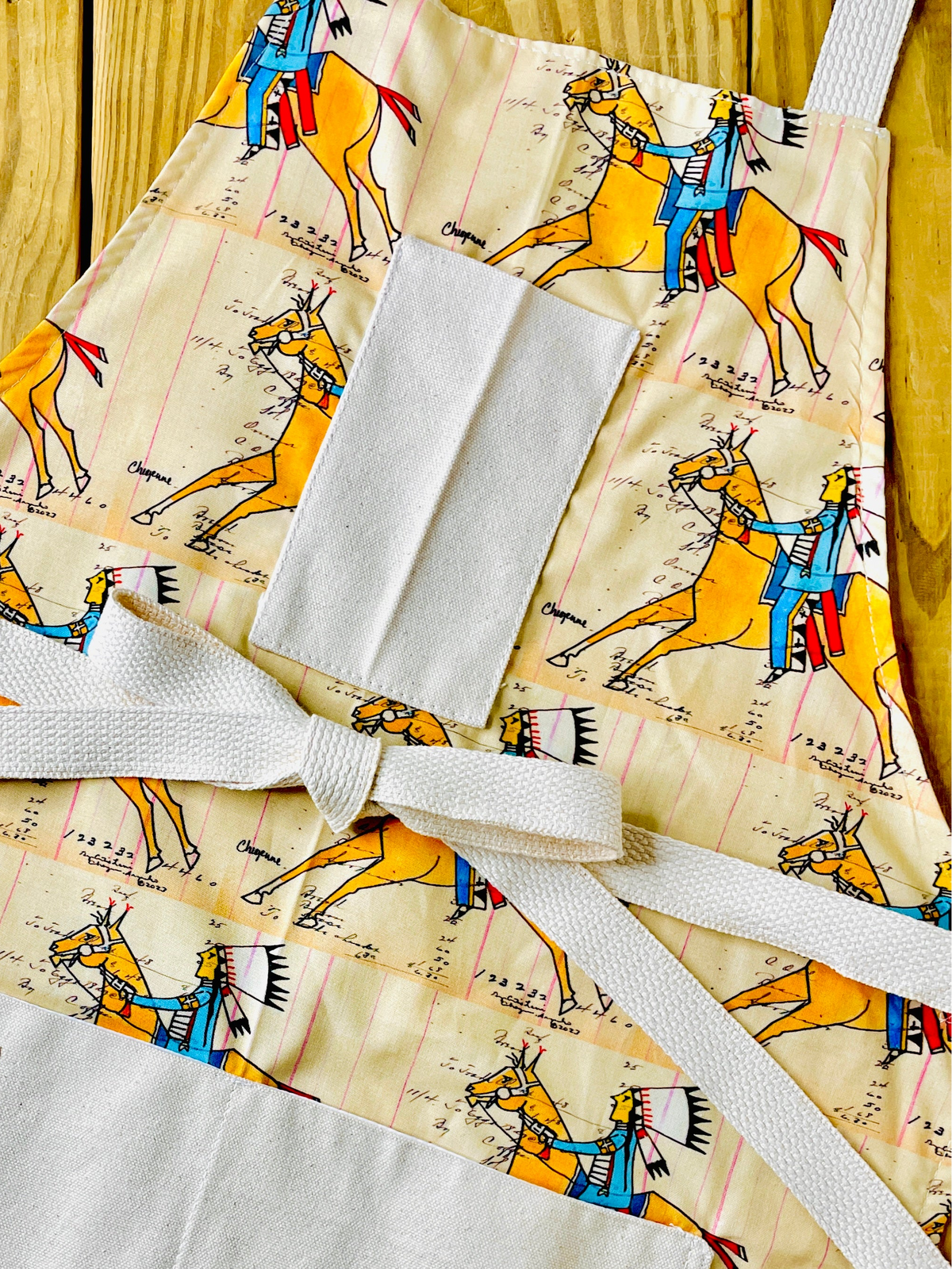 Amanda Bright Designs cheyenne warrior indian chief apron with cream background and a repeating print of indian on horseback wearing red and blue with his headdress on and figural marks on the pattern