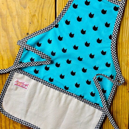 Amanda Bright Designs handmade cotton children’s black cat apron featuring black cat heads and whiskers on a turquoise background with black gingham trim and a 3-pocket canvas panel