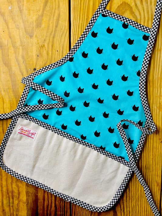 Amanda Bright Designs handmade cotton children’s black cat apron featuring black cat heads and whiskers on a turquoise background with black gingham trim and a 3-pocket canvas panel