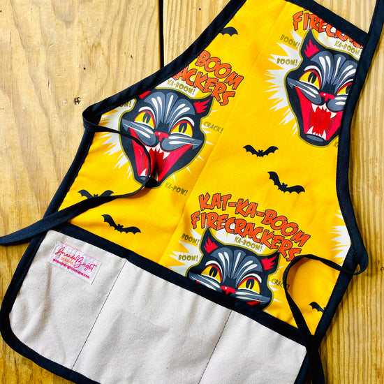 Amanda Bright Designs gold and black cat children’s apron with black cat fireworks screaming cat-face and bats flying. The apron has black trim, natural canvas front pockets, and black neck and waist ties