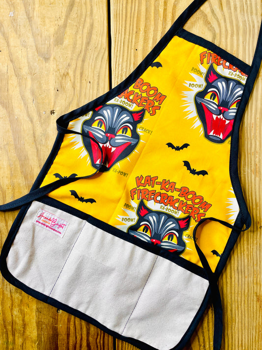 Amanda Bright Designs gold and black cat children’s apron with black cat fireworks screaming cat-face and bats flying. The apron has black trim, natural canvas front pockets, and black neck and waist ties