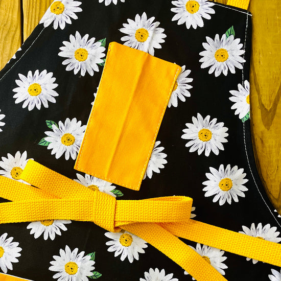 Amanda Bright Designs white daisy with yellow centers pattern apron on black with gold strings and pockets