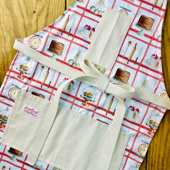 Amanda Bright Designs white baking apron with a red checkered pattern featuring illustrations of baking-related items like a scale, cake, pie, and rolling pin domed cupcakes with canvas pockets and cotton strings