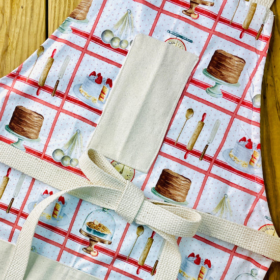 Amanda Bright Designs white baking apron with a red checkered pattern featuring illustrations of baking-related items like a scale, cake, pie, and rolling pin domed cupcakes with canvas pockets and cotton strings