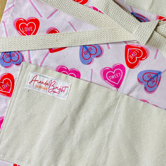 amanda bright designs close-up of a pink cotton apron with a Valentine's Day heart pattern, featuring red, pink, and blue hearts with 'CUPID', 'LOVE', and 'SWEET' printed inside. The apron has a solid beige canvas bib and matching beige twine straps tied in a bow, laid flat on a wooden surface.