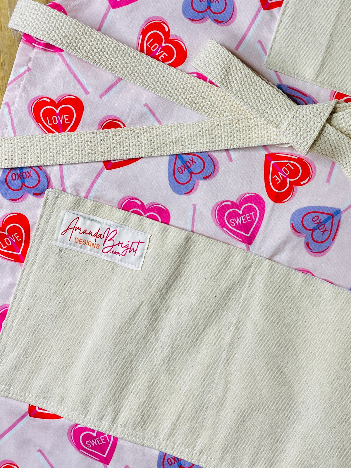 amanda bright designs close-up of a pink cotton apron with a Valentine's Day heart pattern, featuring red, pink, and blue hearts with 'CUPID', 'LOVE', and 'SWEET' printed inside. The apron has a solid beige canvas bib and matching beige twine straps tied in a bow, laid flat on a wooden surface.