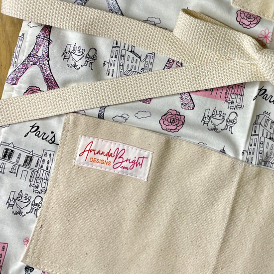 Close-up of apron with a Paris-themed print featuring the Eiffel Tower, buildings, clouds, and pink roses on a cream background. 