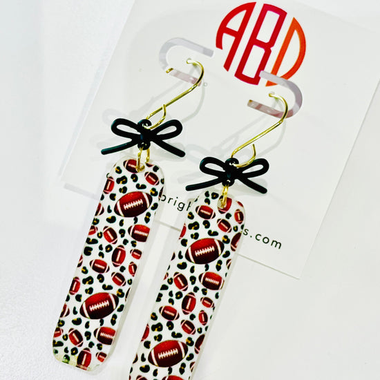 Amanda Bright Designs pair of dangling game day earrings with rectangular shape and football pattern, accented with small black bows and gold hooks