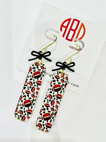 Amanda Bright Designs pair of dangling game day earrings with rectangular shape and football pattern, accented with small black bows and gold hooks