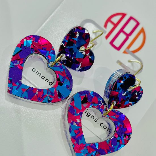 Amanda Bright Designs USA made pair of heart-shaped acrylic dangle earrings with a vibrant multicolor confetti pattern in pink, blue, and purple, featuring gold-tone hooks.