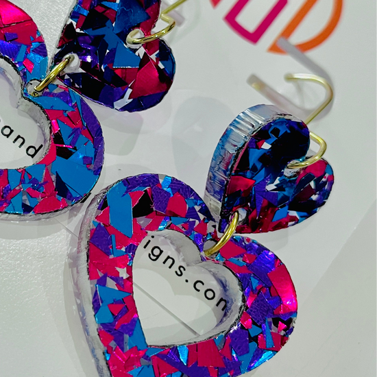 Amanda Bright Designs USA made pair of heart-shaped acrylic dangle earrings with a vibrant multicolor confetti pattern in pink, blue, and purple, featuring gold-tone hooks.