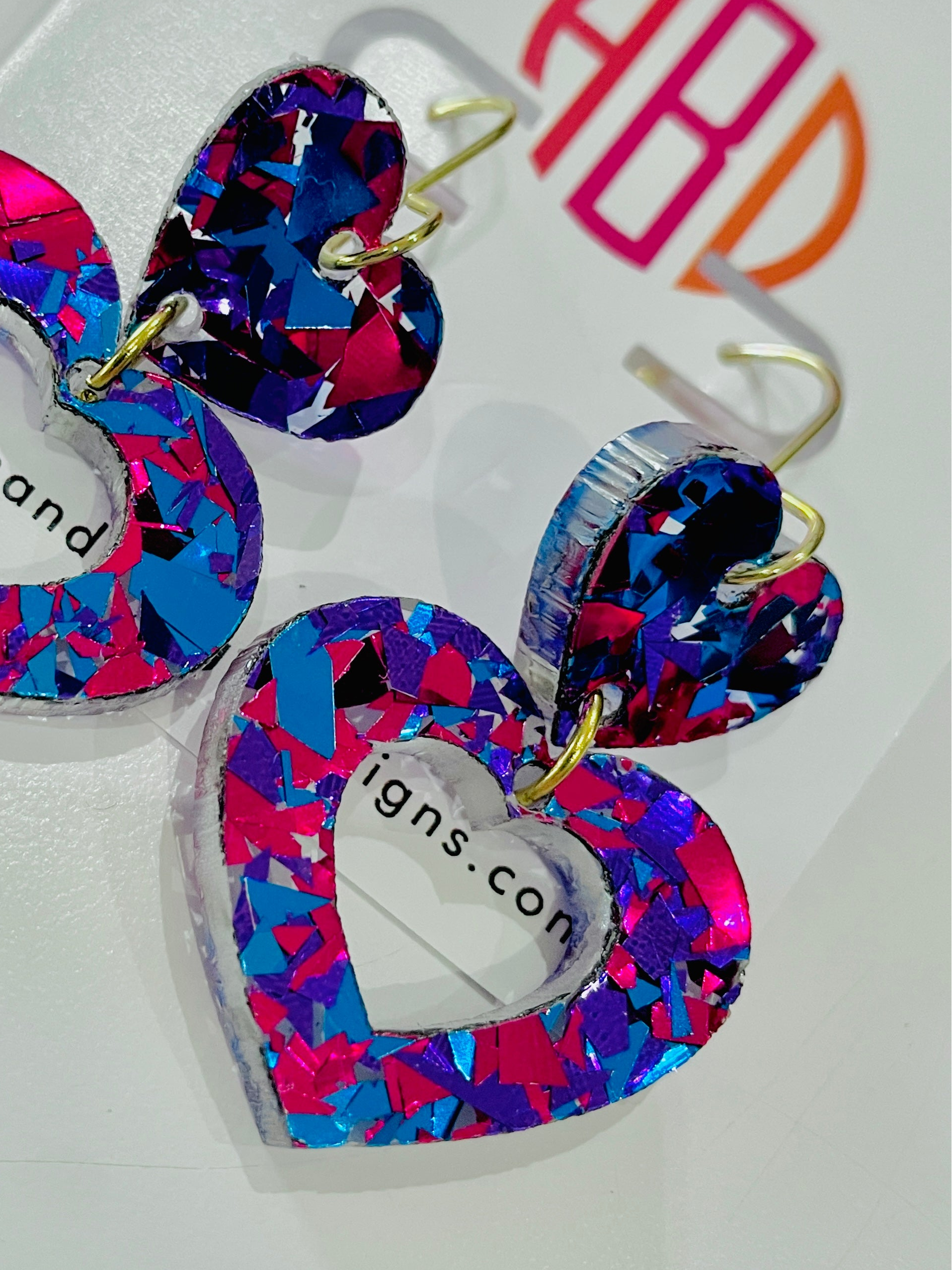 Amanda Bright Designs USA made pair of heart-shaped acrylic dangle earrings with a vibrant multicolor confetti pattern in pink, blue, and purple, featuring gold-tone hooks.