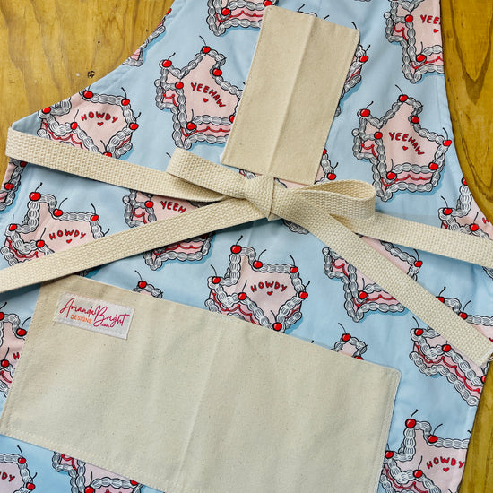 Amanda Bright Designs Howdy Cherry Cowgirl Apron featuring state of texas in cake form with words yeehaw and howdy in icing with cherries on baby blue background