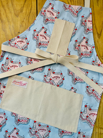 Amanda Bright Designs Howdy Cherry Cowgirl Apron featuring state of texas in cake form with words yeehaw and howdy in icing with cherries on baby blue background