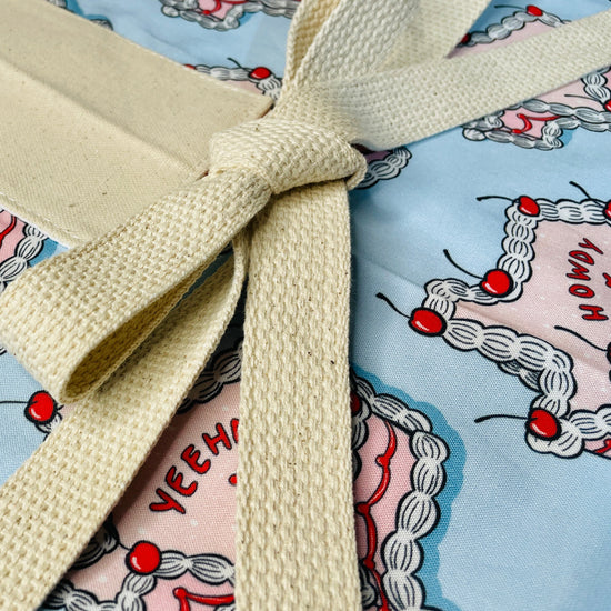 Amanda Bright Designs howdy cherry cowgirl fabric apron texas shaped like a cake with pink icing cherries and words yeehaw and howdy written on the cake on a baby blue background