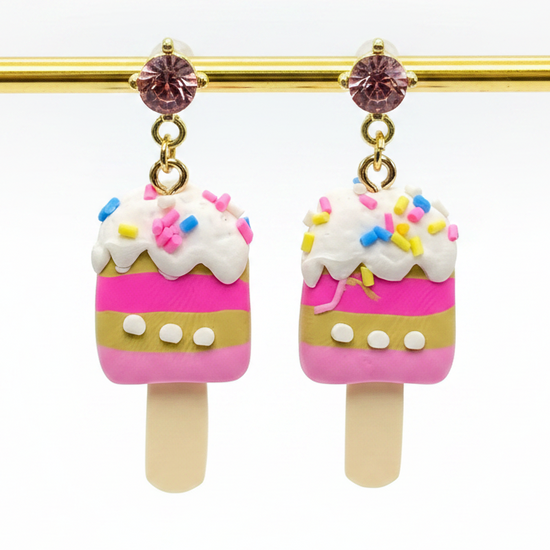 Ice cream pop-shaped earrings with pink and white design on a gold stick against a white background.