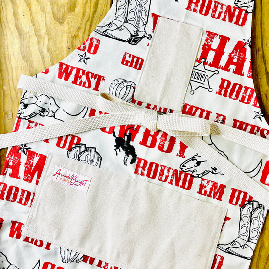 Amanda Bright Designs Cowboy-themed aprons with text and graphics Jane Long Mother of Texas boots, steer, sheriff badge, stars, horseshoes, roping cowboy, horse