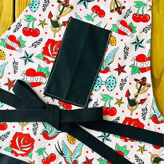 Amanda Bright Designs colorful tattoo-style nautical print apron with cherries, roses, stars, anchors and swallow birds on a blush pink background with black pockets and black strings