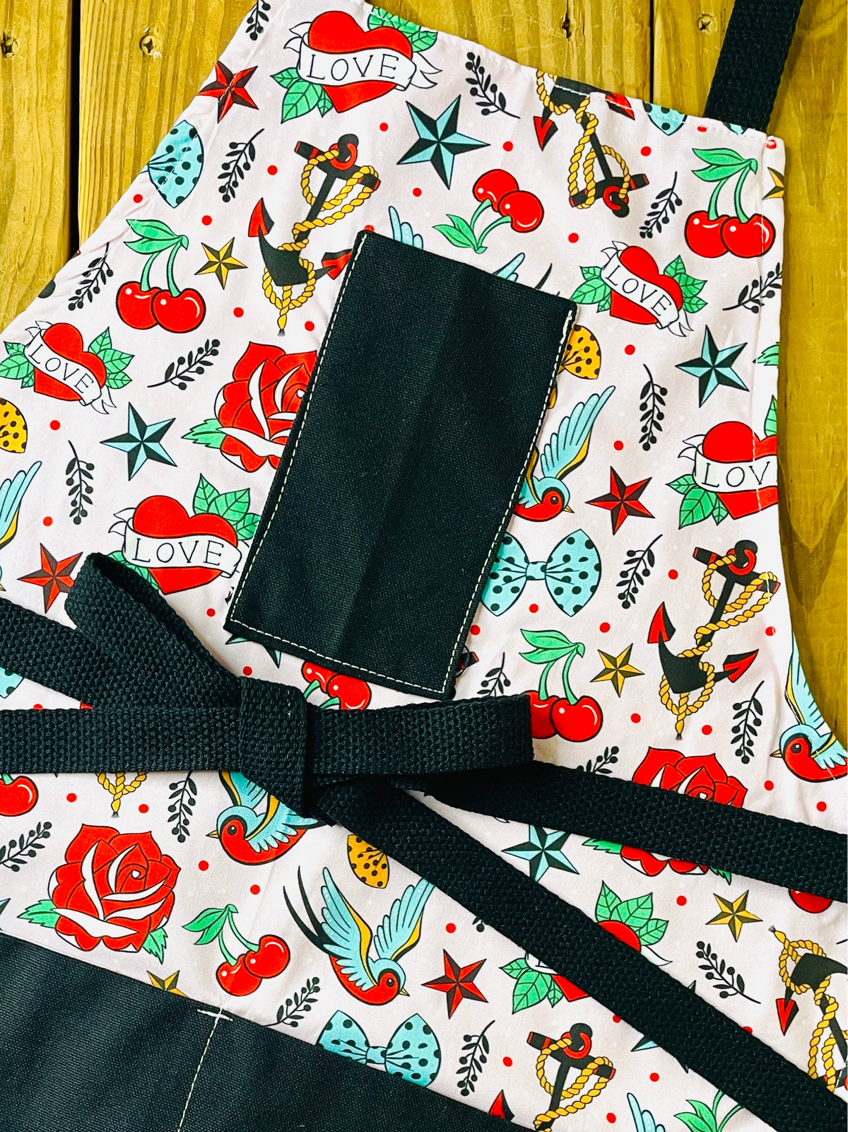 Amanda Bright Designs colorful tattoo-style nautical print apron with cherries, roses, stars, anchors and swallow birds on a blush pink background with black pockets and black strings