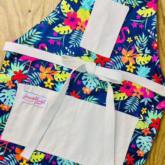 Amanda Bright Designs Nikki multicolored tropical Florida Hawaii floral apron with pink flamingo patterns on a blue background, featuring cotton strings and canvas pockets