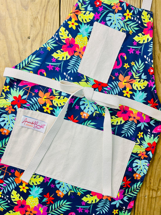 Amanda Bright Designs Nikki multicolored tropical Florida Hawaii floral apron with pink flamingo patterns on a blue background, featuring cotton strings and canvas pockets