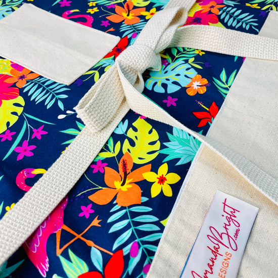 Amanda Bright Designs Nikki multicolored tropical Florida Hawaii floral apron with pink flamingo patterns on a blue background, featuring cotton strings and canvas pockets
