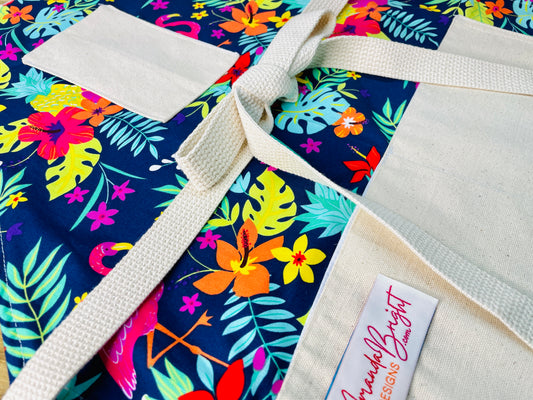 Amanda Bright Designs Nikki multicolored tropical Florida Hawaii floral apron with pink flamingo patterns on a blue background, featuring cotton strings and canvas pockets
