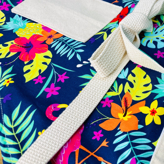 Amanda Bright Designs Nikki multicolored tropical Florida Hawaii floral apron with pink flamingo patterns on a blue background, featuring cotton strings and canvas pockets