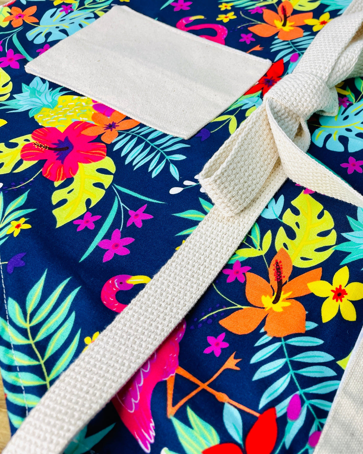 Amanda Bright Designs Nikki multicolored tropical Florida Hawaii floral apron with pink flamingo patterns on a blue background, featuring cotton strings and canvas pockets