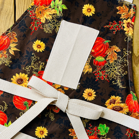 Amanda Bright Designs apron Fabric with autumn-themed design featuring pumpkins, flowers, and leaves on a brown background.