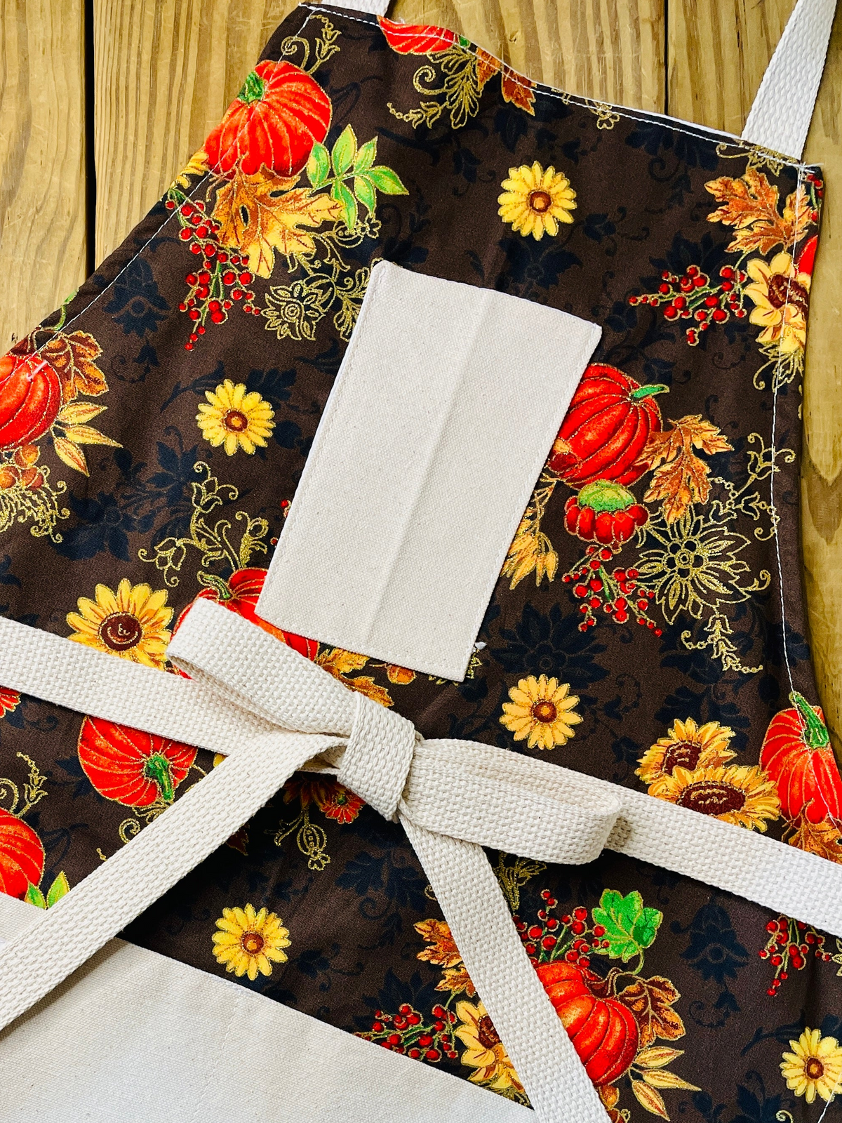Amanda Bright Designs apron Fabric with autumn-themed design featuring pumpkins, flowers, and leaves on a brown background.