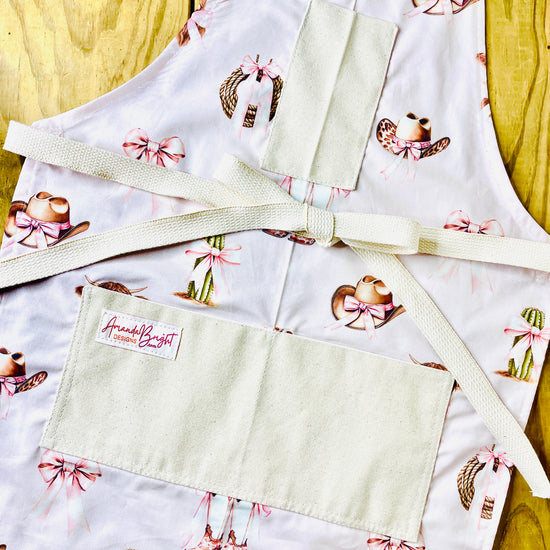 Amanda Bright Designs pink bows highland cow cowboy hat boots cactus and ribbon tied rope on a petal pink background with natural pockets and strings couture apron design