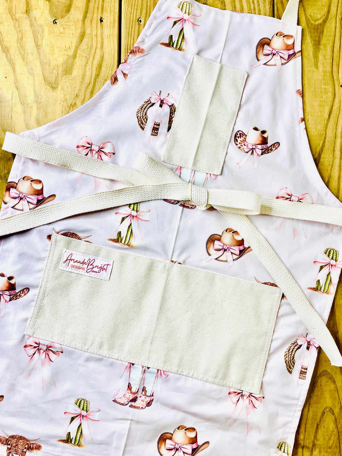 Amanda Bright Designs pink bows highland cow cowboy hat boots cactus and ribbon tied rope on a petal pink background with natural pockets and strings couture apron design