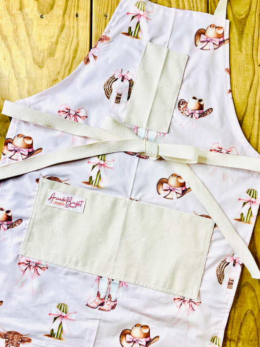 Amanda Bright Designs pink bows highland cow cowboy hat boots cactus and ribbon tied rope on a petal pink background with natural pockets and strings couture apron design