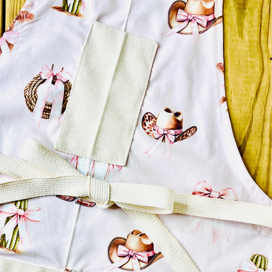 Amanda Bright Designs pink bows highland cow cowboy hat boots cactus and ribbon tied rope on a petal pink background with natural pockets and strings couture apron design