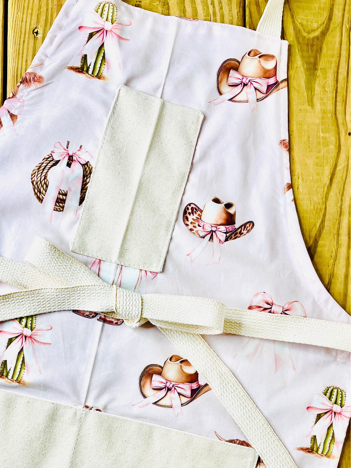 Amanda Bright Designs pink bows highland cow cowboy hat boots cactus and ribbon tied rope on a petal pink background with natural pockets and strings couture apron design