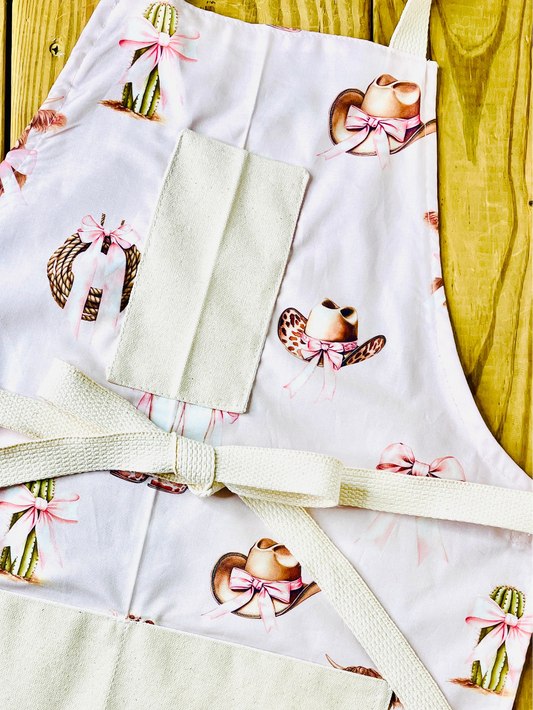 Amanda Bright Designs pink bows highland cow cowboy hat boots cactus and ribbon tied rope on a petal pink background with natural pockets and strings couture apron design